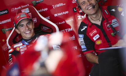 Smiling MotoGP rider in a red racing suit and cap in a sponsor-filled pit with a headset-wearing crew member beside him.