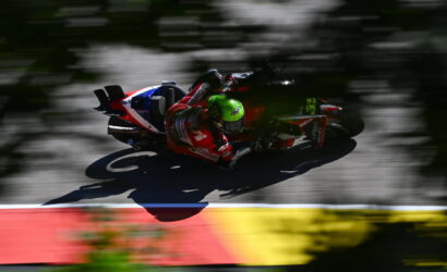 MotoGP rider in red and white gear with a neon green helmet leaning into a turn on a racetrack; motion-blurred background behind the bike.
