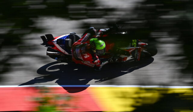 MotoGP rider in red and white gear with a neon green helmet leaning into a turn on a racetrack; motion-blurred background behind the bike.
