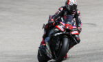 Motorcycle racer in full suit and helmet leaning into a turn on a red-black race bike on a track.