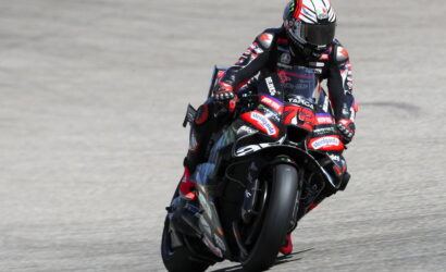 Motorcycle racer in full suit and helmet leaning into a turn on a red-black race bike on a track.