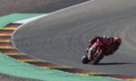 MotoGP rider in red gear leaning into a sharp curve on a race track with painted curbs.