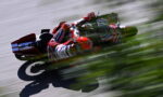 MotoGP rider in red racing suit leans into a turn on a track, sponsor logos visible, with strong motion blur behind.