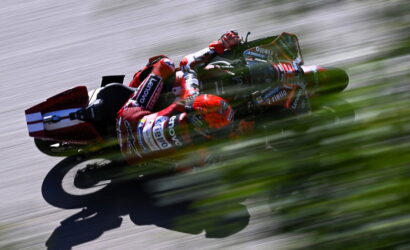 MotoGP rider in red racing suit leans into a turn on a track, sponsor logos visible, with strong motion blur behind.