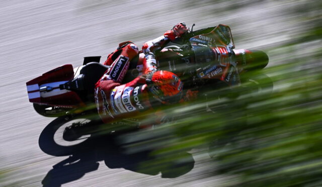 MotoGP rider in red racing suit leans into a turn on a track, sponsor logos visible, with strong motion blur behind.