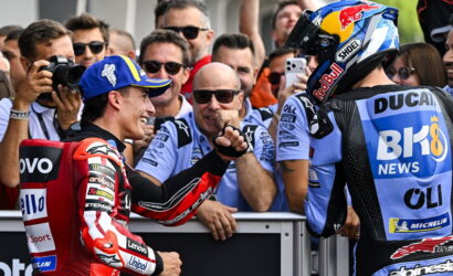 Two MotoGP riders in racing gear share a fist bump amid cheering fans at the track and pit wall behind them in focus.