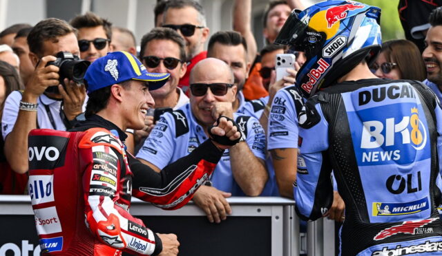 Two MotoGP riders in racing gear share a fist bump amid cheering fans at the track and pit wall behind them in focus.