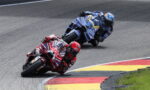 Two motorcycle racers lean into a sharp corner on a racetrack, red bike in front blue bike behind, both in full racing gear.