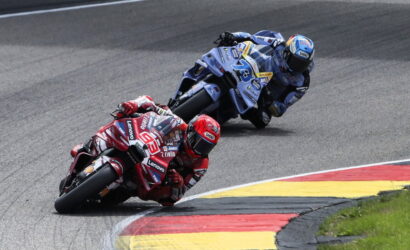 Two motorcycle racers lean into a sharp corner on a racetrack, red bike in front blue bike behind, both in full racing gear.