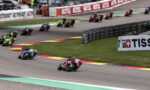 Group of motorcycle racers lean into a corner on a race track, with bright sponsor banners along the barriers in the background.