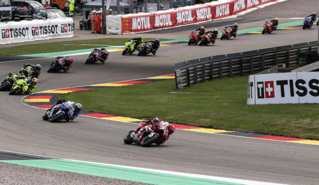 Group of motorcycle racers lean into a corner on a race track, with bright sponsor banners along the barriers in the background.