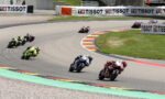 Group of motorcycle racers leaning into a sweeping turn on a race track, red and blue bikes in the foreground with Tissot banners along the barriers.