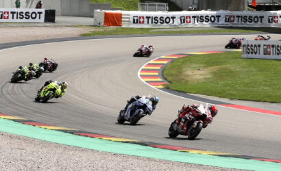 Group of motorcycle racers leaning into a sweeping turn on a race track, red and blue bikes in the foreground with Tissot banners along the barriers.