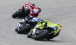 Three sportbike racers lean into a turn on a racetrack; foreground rider wears bright neon yellow gear on a neon-yellow bike with a blue bike behind and a red rider in the rearlust.