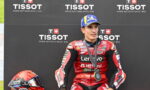 MotoGP rider in red Lenovo racing suit on podium with Tissot backdrop and medals around neck, helmet at left on ground
