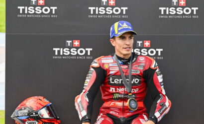 MotoGP rider in red Lenovo racing suit on podium with Tissot backdrop and medals around neck, helmet at left on ground