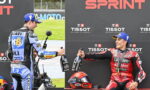 Motorcycle racers on a podium celebrate, each holding a bottle of champagne against a black backdrop with TISSOT logos.
