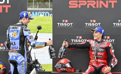 Motorcycle racers on a podium celebrate, each holding a bottle of champagne against a black backdrop with TISSOT logos.