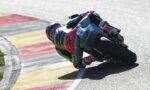MotoGP rider in a colorful racing suit leans into a sharp turn on a race track.