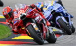MotoGP racers lean into a sharp turn, red bike in front with sponsor logos leading blue bike behind.