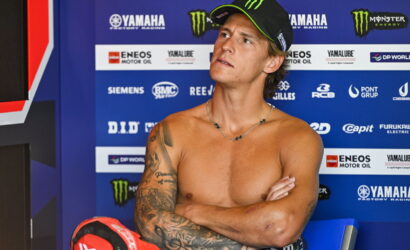 Portrait of a shirtless young man with a tattooed left arm, crossing his arms while wearing a black cap with green trim, seated before a blue Yamaha racing backdrop with sponsor logos.