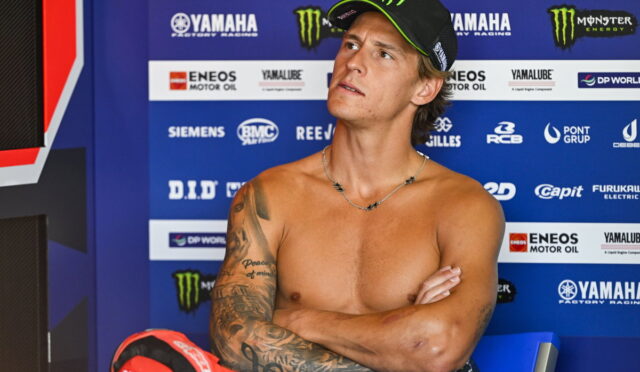 Portrait of a shirtless young man with a tattooed left arm, crossing his arms while wearing a black cap with green trim, seated before a blue Yamaha racing backdrop with sponsor logos.