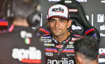 MotoGP rider in an Aprilia Racing cap sits in a racing chair, looking toward the camera during a pit interview.
