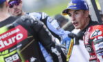 Two motorcycle racers in full gear converse in the paddock; the foreground rider wears a red suit and blue cap facing the camera.