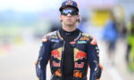 MotoGP rider in a Red Bull KTM racing suit and cap, wearing reflective sunglasses, standing on a race track.