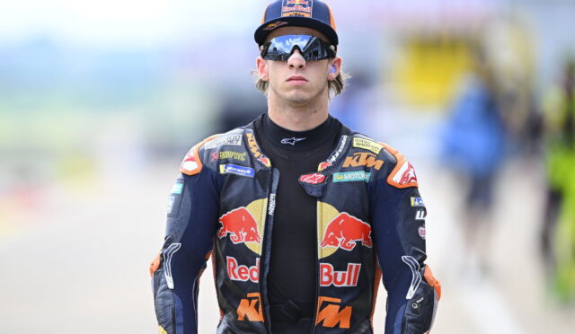 MotoGP rider in a Red Bull KTM racing suit and cap, wearing reflective sunglasses, standing on a race track.