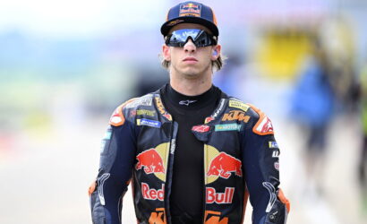 Moto racer in Red Bull KTM suit wearing sunglasses and cap, walking toward the pit area.