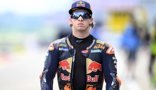 Moto racer in Red Bull KTM suit wearing sunglasses and cap, walking toward the pit area.