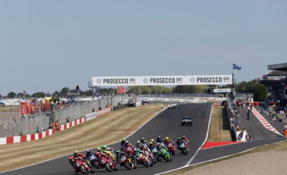 Group of motorcycles racing through a track, riders leaning into a corner as banners read PROSECCO DOC above the course.