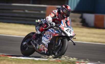 Rider in red and white racing gear leaning into a turn on a race track, sport bike with Union Jack livery and sponsor decals in motion on a sunny day.