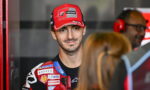 Portrait of a motorcycle racer in a red Ducati Lenovo cap and sponsor-filled racing suit, standing in a garage with crew in the background.