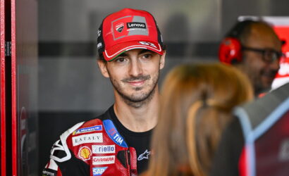 Portrait of a motorcycle racer in a red Ducati Lenovo cap and sponsor-filled racing suit, standing in a garage with crew in the background.