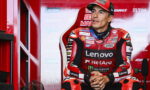 MotoGP rider in a red Lenovo NetApp racing suit sits in a red pit-area, hands clasped, looking off to the side, ready for action/ interview.
