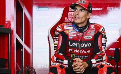 MotoGP rider in a red Lenovo NetApp racing suit sits in a red pit-area, hands clasped, looking off to the side, ready for action/ interview.