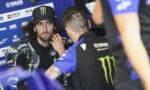 MotoGP rider with Monster Energy cap sits in a blue Yamaha paddock, listening to a team member.