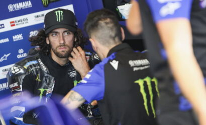 MotoGP rider with Monster Energy cap sits in a blue Yamaha paddock, listening to a team member.