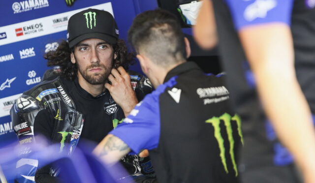 MotoGP rider with Monster Energy cap sits in a blue Yamaha paddock, listening to a team member.
