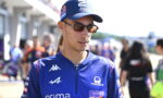 Male cyclist in blue racing kit wearing sunglasses and a cap, standing in a busy pit/paddock area at a race event.