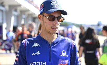 Male cyclist in blue racing kit wearing sunglasses and a cap, standing in a busy pit/paddock area at a race event.