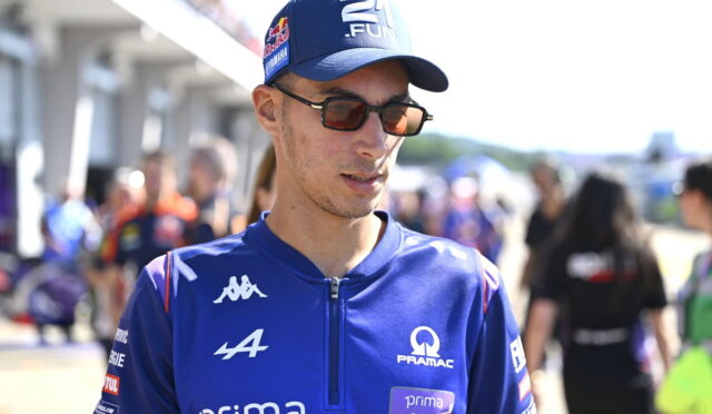 Male cyclist in blue racing kit wearing sunglasses and a cap, standing in a busy pit/paddock area at a race event.