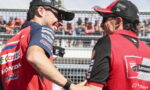 Two motorcycle racers in red team gear shake hands and smile at a race event, spectators behind a fence in the background.