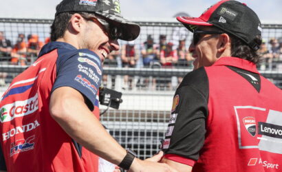 Two motorcycle racers in red team gear shake hands and smile at a race event, spectators behind a fence in the background.