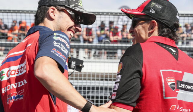 Two motorcycle racers in red team gear shake hands and smile at a race event, spectators behind a fence in the background.