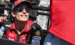 Male race driver in a red team shirt and cap, wearing dark sunglasses, smiling in a pit area with sponsorship logos visible on the jersey.