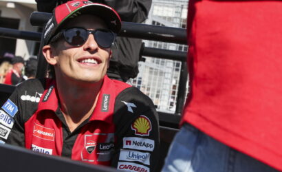 Male race driver in a red team shirt and cap, wearing dark sunglasses, smiling in a pit area with sponsorship logos visible on the jersey.
