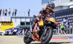 Moto racer on orange-red KTM bike numbered 25 doing a sharp turn on a race track; Red Bull branding visible, spectators in background.